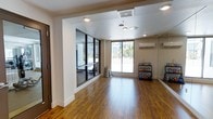 This single-floor fitness facility is designed to support a variety of workout routines in a clean, functional environment. The main exercise area features warm hardwood flooring that extends throughout the space, creating an inviting and energetic atmosphere. Large sliding glass doors allow abundant natural light to fill the room while offering views of the outdoor surroundings, contributing to an open and airy feel. A wall-mounted mini-split HVAC unit ensures year-round climate comfort during workouts. Recessed lighting and flush-mount ceiling fixtures provide consistent illumination throughout. A dedicated kettlebell rack stocked with color-coded weights of varying sizes is conveniently positioned near the exterior wall, making equipment easily accessible. One side of the space features a glass-enclosed room framed in sleek black metal, visible through floor-to-ceiling glass panels — ideal for group classes, personal training sessions, or specialized equipment use. Mirrored wall sections enhance the sense of space and support form-checking during exercise. The overall design is modern and minimalist, with neutral wall tones and clean architectural lines that keep the focus on functionality and movement.