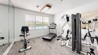 Located at 100 Marshview Dr in Newport News, Virginia, this dedicated exercise room offers a well-organized fitness space designed to support a variety of workout routines. The room features neutral-toned carpet flooring and crisp white walls, creating a clean and functional environment. Large windows fitted with blinds allow natural light to fill the space, with views of what appears to be a pool area just outside, adding a refreshing outdoor connection.

The room is equipped with a diverse selection of fitness equipment, including a treadmill, a Matrix recumbent exercise bike, a multi-station cable strength-training machine, and additional cardio equipment. A medicine ball rack stocked with a colorful assortment of weighted balls provides options for functional training, while workout reference charts — covering bodyweight exercises, yoga poses, and medicine ball routines — are posted on the wall for user guidance.

A full-length mirror along one wall enhances the sense of space and allows users to monitor their form during exercise. Overhead, warm-toned wooden light fixtures provide supplemental illumination. The room also includes a seating area with a chair, offering a convenient rest space. This thoughtfully arranged fitness room delivers a comprehensive workout experience in a compact, well-lit setting.