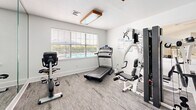 Located at 100 Marshview Dr in Newport News, Virginia, this dedicated exercise room offers a well-organized fitness space designed to support a variety of workout routines. The room features neutral-toned carpet flooring and crisp white walls, creating a clean and functional environment. Large windows fitted with blinds allow natural light to fill the space, with views of what appears to be a pool area just outside, adding a refreshing outdoor connection.

The room is equipped with a diverse selection of fitness equipment, including a treadmill, a Matrix recumbent exercise bike, a multi-station cable strength-training machine, and additional cardio equipment. A medicine ball rack stocked with a colorful assortment of weighted balls provides options for functional training, while workout reference charts — covering bodyweight exercises, yoga poses, and medicine ball routines — are posted on the wall for user guidance.

A full-length mirror along one wall enhances the sense of space and allows users to monitor their form during exercise. Overhead, warm-toned wooden light fixtures provide supplemental illumination. The room also includes a seating area with a chair, offering a convenient rest space. This thoughtfully arranged fitness room delivers a comprehensive workout experience in a compact, well-lit setting.
