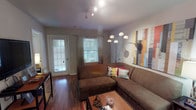 This single-floor residence is thoughtfully designed with an open, flowing layout that connects the main living areas seamlessly. The living room serves as a welcoming central space, featuring rich hardwood-style flooring, track lighting overhead, and a large sectional sofa positioned around a coffee table. Two windows allow natural light to filter in, and a glazed exterior door provides additional brightness and outdoor access. Wall art and a multi-arm arc floor lamp add decorative character to the room.

The living area opens directly into a dedicated dining space, where pendant lighting hangs above a round dining table with chairs. This transitions naturally into the galley-style kitchen, which is well-equipped with warm wood cabinetry, grey countertops, black appliances including a range and over-the-range microwave, and a double-basin sink. The kitchen offers ample storage and counter workspace along both sides.

The bedroom is carpeted for comfort and features a window dressed with decorative curtains, a reach-in closet with sliding doors, and space for a desk and dresser. The bathroom is efficiently laid out with a wood-vanity cabinet, a single-basin sink, a lighted mirror, and a shower-tub combination.