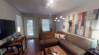 This single-floor residence is thoughtfully designed with an open, flowing layout that connects the main living areas seamlessly. The living room serves as a welcoming central space, featuring rich hardwood-style flooring, track lighting overhead, and a large sectional sofa positioned around a coffee table. Two windows allow natural light to filter in, and a glazed exterior door provides additional brightness and outdoor access. Wall art and a multi-arm arc floor lamp add decorative character to the room.

The living area opens directly into a dedicated dining space, where pendant lighting hangs above a round dining table with chairs. This transitions naturally into the galley-style kitchen, which is well-equipped with warm wood cabinetry, grey countertops, black appliances including a range and over-the-range microwave, and a double-basin sink. The kitchen offers ample storage and counter workspace along both sides.

The bedroom is carpeted for comfort and features a window dressed with decorative curtains, a reach-in closet with sliding doors, and space for a desk and dresser. The bathroom is efficiently laid out with a wood-vanity cabinet, a single-basin sink, a lighted mirror, and a shower-tub combination.