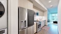 This thoughtfully designed single-floor residence showcases a clean, contemporary aesthetic with cohesive finishes throughout. The galley-style kitchen is a functional highlight, featuring stainless steel appliances including a gas range, over-the-range microwave, dishwasher, and a large French door refrigerator. Light wood-grain cabinetry extends floor to ceiling, complemented by a white subway tile backsplash and crisp white countertops, creating a bright and efficient cooking space. Hardwood-style flooring runs seamlessly through the kitchen and into the main living areas, adding warmth to the modern design. The living room is anchored by a large picture window that floods the space with natural light and offers views of the surrounding urban streetscape. A textured accent wall adds visual depth and character to the room. A dedicated dining nook with a compact bistro table provides a charming spot for casual meals. The bedroom is serene and minimalist, with crisp white walls and access to a well-appointed walk-in closet with custom wood shelving. A sliding barn-style door connects the bedroom to the living area. The full bathroom is finished with large-format white tile flooring, a soaking tub with shower, and a light wood vanity, maintaining the residence's clean and modern palette throughout.