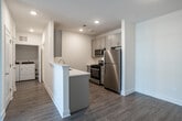 Located at 1200 Felix Way in Westampton, NJ, this single-floor 2-bedroom, 1-bathroom residence offers a clean, modern aesthetic with thoughtfully selected finishes throughout. The open-concept kitchen and living area serves as the heart of the home, featuring a peninsula with a built-in sink, crisp white countertops, gray shaker-style cabinetry, stainless steel appliances including a gas range and microwave, and warm wood-look flooring. The living area extends naturally from the kitchen and is bathed in natural light through sliding glass doors that open to an outdoor view. Both bedrooms are carpeted for comfort and maintain a bright, neutral palette. The primary bedroom offers generous proportions and includes a spacious walk-in closet with wire shelving and hanging rods for ample storage. The second bedroom features a large window that fills the room with natural light. The full bathroom is finished with elegant marble-look tile flooring, a gray vanity with sleek hardware, a large mirror with overhead lighting, and a full tub-shower combination. An in-unit washer and dryer area is also present, adding everyday convenience. Throughout, recessed lighting, crisp white walls, and clean trim work contribute to a cohesive and move-in-ready living environment.