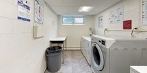 This shared laundry facility is a well-organized, single-floor utility space designed to accommodate the everyday laundry needs of building residents. The room features a combination of commercial-grade top-loading and front-loading washers alongside matching dryers, providing ample capacity for regular use. A dedicated utility sink offers added convenience for pre-treating garments or handwashing delicate items.

A sturdy folding table provides a flat surface for sorting and folding laundry, keeping the workflow efficient and tidy. Posted instructional guides for washing and drying are displayed on the walls, making the space accessible and easy to use for all residents. A waste bin is conveniently positioned within the room for easy disposal.

The room is finished with painted concrete block walls and light tile flooring that is easy to maintain. A drop ceiling with recessed fluorescent lighting ensures the space is well-lit throughout. A small ground-level window allows for a degree of natural light and ventilation. Access is provided through a solid metal door, and exposed utility piping and dryer venting are neatly routed along the walls. Overall, the space is clean, practical, and thoughtfully laid out to support efficient shared use.