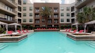This outdoor pool area offers a well-appointed resort-style retreat nestled within the courtyard of a modern multi-story building. The centerpiece is a generously sized swimming pool with clear turquoise water, framed by a smooth tile deck and surrounded by rows of red lounge chairs ideal for sunbathing and relaxation. A central raised planter with lush greenery and ornamental grasses adds a natural focal point between the pool and the building facade. Towering palm trees punctuate the space, lending a tropical ambiance to the outdoor setting. Two striped black-and-white cabanas serve as private lounging retreats, each equipped with comfortable wicker sectional seating, accent pillows, mounted flat-screen televisions, and curtained sides for shade and privacy. Manicured hedgerows, decorative planters, and wrought iron fencing contribute to the polished, landscaped aesthetic throughout. The surrounding multi-story building, finished in a combination of brick and neutral stucco with large windows and private balconies, encloses the courtyard on multiple sides, creating a sense of seclusion. The tile pool deck provides ample open space for lounging, while the overall design balances modern architecture with inviting outdoor comfort.