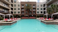 This outdoor pool area offers a well-appointed resort-style retreat nestled within the courtyard of a modern multi-story building. The centerpiece is a generously sized swimming pool with clear turquoise water, framed by a smooth tile deck and surrounded by rows of red lounge chairs ideal for sunbathing and relaxation. A central raised planter with lush greenery and ornamental grasses adds a natural focal point between the pool and the building facade. Towering palm trees punctuate the space, lending a tropical ambiance to the outdoor setting. Two striped black-and-white cabanas serve as private lounging retreats, each equipped with comfortable wicker sectional seating, accent pillows, mounted flat-screen televisions, and curtained sides for shade and privacy. Manicured hedgerows, decorative planters, and wrought iron fencing contribute to the polished, landscaped aesthetic throughout. The surrounding multi-story building, finished in a combination of brick and neutral stucco with large windows and private balconies, encloses the courtyard on multiple sides, creating a sense of seclusion. The tile pool deck provides ample open space for lounging, while the overall design balances modern architecture with inviting outdoor comfort.