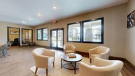 This single-floor community lounge and leasing space offers a well-appointed, open-plan layout designed to serve as both a social hub and functional workspace. The interior is finished with warm-toned wood-look flooring throughout, recessed lighting, and a ceiling fan that contributes to a comfortable, airy atmosphere.

The lounge area is anchored by a seating arrangement of cream upholstered chairs grouped around a round coffee table atop a textured area rug, creating an inviting space for conversation and relaxation. A wall-mounted flat-screen TV and large-scale decorative artwork add visual interest to the space.

Along one wall, a kitchenette features teal cabinetry paired with a butcher-block countertop island, a stainless steel refrigerator, and beverage station — offering convenient amenities for residents and guests. An adjacent bistro-height table with industrial-style stools provides a casual dining or work option.

A dedicated workstation alcove, finished with textured walls and a built-in granite-top desk, provides a quiet area suited for individual productivity. 

The space is lined with large, black-framed windows and double glass doors that flood the interior with natural light while offering views of the surrounding residential community. The overall design balances modern aesthetics with functional comfort.