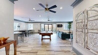 Located at 3450 Miller Dr in Chamblee, Georgia, this single-floor community clubhouse is thoughtfully designed to serve as a social and recreational hub. The open layout is anchored by light wood-look flooring that runs throughout, complemented by sage green crown molding and recessed lighting that create a cohesive, contemporary aesthetic.

The central gathering area features a full-size pool table as its focal point, surrounded by ample open space. Multiple ceiling fans overhead ensure comfortable airflow throughout. Large sliding glass doors lead to an outdoor balcony, filling the interior with natural light. Adjacent to this main area is a dedicated game nook housing a shuffleboard table, high-top tables, and bar-height seating, with decorative lattice wall panels adding visual interest.

The fully equipped kitchen is a standout feature, showcasing gray shaker-style cabinetry, granite countertops, a glossy white subway tile backsplash, stainless steel appliances including a range with a wall-mounted hood, and a generous center island with bar seating. An industrial-style pendant light fixture adds warmth overhead.

Separate lounge zones provide quieter retreat spaces, each furnished with plush sofas, coffee tables, wall-mounted televisions, and expansive windows that bring in abundant natural light, completing this well-appointed communal space.