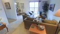 This single-floor residence features an open-concept layout that seamlessly connects the living and dining areas, creating a cohesive and functional flow. The living room is generously proportioned, accommodating a full sofa set arranged around a warm wood-toned coffee table. Decorative touches, including potted orchids, a small indoor tree, and tasteful framed artwork on the walls, contribute to a welcoming ambiance. Neutral wall-to-wall carpeting runs throughout, adding warmth underfoot and unifying the space visually.

A large sliding glass door dominates one wall of the living area, flooding the room with natural light and providing direct access to an outdoor balcony. The balcony extends the living space outward, offering an area for outdoor relaxation. Framed by curtain panels, the door serves as both a functional and aesthetic focal point.

Adjacent to the living room, the dining area is visible through an open passageway and features a wood dining set beneath a chandelier-style light fixture, creating a defined yet connected dining space. The overall layout is practical and inviting, suited for everyday living and casual entertaining.