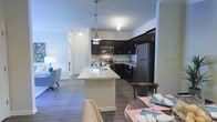 This single-floor residence offers a thoughtfully designed open-concept layout that seamlessly connects the kitchen, dining, and living areas into one cohesive, flowing space. The kitchen is a standout feature, showcasing rich dark cabinetry, granite-look countertops, and a central island that provides additional prep space and casual seating. Stainless steel appliances — including a refrigerator, microwave, and range — add a sleek, modern touch, while stylish pendant lighting overhead enhances the overall ambiance. Wood-look plank flooring runs throughout the main living areas, adding warmth and a sense of continuity. The dining area sits adjacent to the kitchen, creating a natural gathering space ideal for entertaining. The living area, partially visible from the kitchen, is bright and inviting, with soft, neutral tones that contribute to a calm, welcoming atmosphere. A hallway leads to additional rooms, suggesting a well-organized and functional floor plan. Track lighting and recessed fixtures ensure the space is well-lit throughout. Overall, this residence balances contemporary design with practical livability, making it an attractive and comfortable single-floor home.