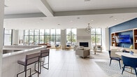 Located at 1178 Silas Deane Highway in Wethersfield, Connecticut, this single-floor community amenity space is designed with a sleek, contemporary aesthetic that balances social gathering and recreational activity. The open-concept layout is anchored by expansive floor-to-ceiling windows that flood the interior with natural light and offer views of the surrounding greenery and outdoor parking areas.

The lounge area features a sophisticated arrangement of neutral-toned sofas and accent chairs centered around a modern linear fireplace, accented by geometric pendant lighting and large-format white tile flooring throughout. Bold navy-blue textured accent walls provide a striking contrast to the bright, airy atmosphere, lending the space a refined yet welcoming character.

A built-in bar with counter seating serves as a social hub, equipped with appliances and ample prep space, and is complemented by a mounted flat-screen television. Adjacent to the bar, a dedicated game room area houses a billiards table and a shuffleboard table, ideal for recreational use. A dining zone with round tables and blue accent chairs offers a casual setting for meals or group gatherings. Geometric pendant fixtures and recessed lighting add layers of illumination throughout, enhancing the space's modern design. The overall ambiance is vibrant, polished, and well-suited for community engagement.