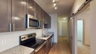 This 1-bedroom, 1-bathroom unit showcases a thoughtfully designed interior with modern finishes throughout. The kitchen is a standout feature, equipped with stainless steel appliances including an electric range, built-in microwave, and dishwasher. Rich dark-toned cabinetry with sleek hardware lines the walls, complemented by granite countertops and a white subway tile backsplash. A stainless steel undermount sink with a contemporary faucet adds a polished touch. Track lighting overhead provides focused illumination across the kitchen workspace. Warm-toned hardwood-style flooring runs throughout the unit, lending a cohesive and inviting feel. The hallway leading from the kitchen connects the spaces efficiently, with a sliding barn door adding a stylish architectural detail. The overall design balances functionality with contemporary aesthetics, making use of clean lines, neutral tones, and quality materials. The single-floor layout ensures easy navigation between spaces, and the well-considered finishes throughout give the unit a modern, move-in-ready appeal suited for comfortable everyday living.