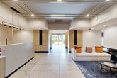 Located at 50 Park Row W in Providence, Rhode Island, this expansive amenity floor offers a thoughtfully designed and elegantly appointed communal space. The grand entrance lobby immediately impresses with large-format marble-look tile flooring, a staffed reception desk finished in warm wood and stone tones, and sophisticated wall paneling in light wood with dark accents. Recessed lighting and structured ceiling details lend a polished, contemporary feel throughout. Wide, light-filled hallways connect the various areas, accented by warm tile flooring, cream-toned paneling, and lush indoor greenery. The combined kitchen and living room area is bright and airy, featuring floor-to-ceiling windows that frame lush outdoor views of Providence's skyline and a landscaped terrace. This space is furnished with designer seating, a marble-top coffee table, a statement chandelier, and a dining area beneath a sleek linear pendant light. A dedicated activity room provides a large gathering table, comfortable seating, and built-in storage with games and leisure supplies. The kitchen nook features dark cabinetry with gold hardware, white quartz countertops, and textured tile backsplash. Outside, a beautifully manicured courtyard garden with winding pathways, vibrant plantings, and a raised outdoor terrace with lounge seating completes this exceptional amenity offering.