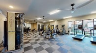 This spacious fitness center is designed to accommodate a comprehensive range of workout routines, offering an extensive selection of both cardio and strength training equipment. The layout is thoughtfully organized, with cardio machines—including multiple treadmills, ellipticals, and a stationary bike—positioned along a wall of floor-to-ceiling windows that frame views of the outdoor surroundings, allowing natural light to fill the space during workouts. The strength training area is equally well-appointed, featuring a multi-station cable machine, a Smith machine, a weight bench, and a full rack of dumbbells neatly arranged along mirrored walls. The mirrors visually expand the room and allow users to monitor their form throughout their session. The flooring throughout is a distinctive checkered tile pattern in neutral tones, adding a polished aesthetic to the functional environment. Ceiling fans are mounted overhead and work in tandem with fluorescent strip lighting to maintain a comfortable, well-lit atmosphere. Additional utility doors provide access to what appear to be storage or utility areas. A wall-mounted television is also present, adding entertainment options for users. The entrance features glass-paneled doors with views into a neighboring common area.