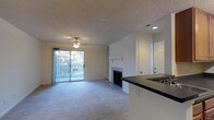 This single-floor residence offers a comfortable and functional layout designed for everyday living. The main living area is a generous, open space finished with soft wall-to-wall carpeting and a textured ceiling. A ceiling fan provides year-round comfort, while a sliding glass door floods the room with natural light and opens directly onto a private deck overlooking lush, mature trees — creating a serene connection to the outdoors.

A tile-surround fireplace serves as a central focal point in the living area, flanked by built-in shelving that provides both display and storage options. The space transitions smoothly into the kitchen area, which features a dark-toned countertop bar that creates a natural boundary between the living and food preparation zones, ideal for casual dining or entertaining.

Throughout the main living space, the neutral color palette of soft whites and warm beiges creates a calm, inviting atmosphere. The layout is straightforward and practical, with interior doors providing access to additional areas of the home. Overall, this residence combines comfortable finishes, practical built-ins, and pleasant outdoor access to offer a well-rounded single-floor living experience.