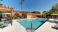 Located at 120 S Cranford Rd in Goose Creek, South Carolina, this outdoor pool facility offers a well-maintained communal amenity space designed for relaxation and recreation. The centerpiece is a generously sized rectangular swimming pool with clearly marked depth indicators, ranging from a shallow entry point to a deeper end, all accessible via chrome handrails. The pool's clear turquoise water reflects the abundant South Carolina sunshine, creating an inviting atmosphere throughout the day.

Surrounding the pool is a broad concrete sundeck furnished with multiple lounge chairs and a shaded seating area featuring a vibrant green umbrella and table-and-chair set, ideal for outdoor gatherings. The entire pool area is enclosed by a sturdy black iron fence, providing both safety and a defined boundary to the space.

The backdrop includes classic brick buildings that frame the area, complemented by mature landscaping of tall pine trees, lush palms, and subtropical greenery that lend the space a distinctly Southern coastal character. A dedicated brick pool house structure with distinctive yellow-green doors provides convenient access to restrooms and storage facilities. The combination of durable brick construction, mature tree canopy, and thoughtful amenities makes this pool area a standout outdoor feature of the Goose Creek property.