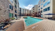 This residential community's amenity level is designed around a central open-air courtyard that serves as the focal point of the space. At its heart lies a sparkling resort-style swimming pool, surrounded by a broad brick-paver deck lined with sleek lounge chairs and shaded dining areas with market umbrellas — ideal for relaxing or socializing outdoors. The multi-story building facades enclosing the courtyard combine warm red brick with modern stucco panels, creating an inviting urban aesthetic punctuated by balconies and large windows throughout.

Landscaped planting beds with ornamental grasses, shrubs, and trees add natural texture and greenery throughout the courtyard, softening the hardscape and enhancing the overall ambiance. A covered outdoor lounge area offers a casual gathering space equipped with wicker furniture, ceiling fans, and a wall-mounted television, making it well-suited for group entertainment. Nearby, a covered game area features a pool table alongside additional seating.

Connecting passageways between buildings are appointed with decorative blue ceramic planters, bench seating, and subtle lighting, ensuring a polished and cohesive aesthetic throughout. The entry features glass double doors set within a brick-and-stucco surround, presenting a clean and professional welcome to all who enter.
