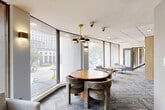 Located at 8750 Georgia Ave in Silver Spring, Maryland, this single-floor amenity space offers a well-appointed community lounge designed for both recreation and relaxation. The layout follows a gently curving footprint, creating a dynamic flow between distinct activity zones, all unified by patterned carpeting and a drop tile ceiling with recessed and pendant lighting throughout.

The lounge area features a curved glass exterior wall that floods the space with natural light and provides views of the surrounding streetscape and greenery. Comfortable seating arrangements — including upholstered lounge chairs and accent tables — are positioned to take advantage of the panoramic windows. A round wood dining table with sculptural upholstered chairs anchors the dining area beneath a gold-toned multi-arm pendant light.

The game room is a standout feature, housing a full-size billiards table beneath a bold cluster pendant fixture, alongside wire-frame bistro tables and chairs set against the expansive window wall. Additional recreational amenities include a foosball table, a bar-height table with seating, and wall-mounted artwork that adds a contemporary touch. A private media room offers a large flat-screen TV, a wood conference table, and lush potted plants. Cue storage and a small kitchenette area provide practical support for the game room. The overall ambiance is modern, social, and thoughtfully designed for community engagement.