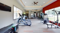 Located at 3130 Hillandale Rd in Durham, NC, this well-appointed fitness center offers a thoughtfully designed space for residents to pursue their health and wellness goals. The single-floor layout is organized to accommodate a full range of exercise activities, from cardio to strength training.

The space features elegant tray ceilings with warm recessed lighting and ceiling fans, creating a comfortable and inviting atmosphere. Neutral gray walls are accented by bold red feature walls, adding a dynamic, energetic character to the interior. The patterned carpet flooring with orange accent stripes adds a modern touch throughout.

Cardio equipment is a highlight, with multiple treadmills, elliptical machines, and a stationary bike — each equipped with individual digital screens for an engaging workout experience. The strength training area includes a cable machine, adjustable weight bench, a full dumbbell rack, and accessory equipment such as battle ropes and medicine balls.

Large windows along multiple walls flood the space with natural light and offer views of the surrounding greenery, enhancing the workout environment. Wall-mounted televisions are positioned throughout for entertainment. A water station with a red mosaic tile accent wall provides a stylish and functional hydration point. Direct exterior door access adds convenience for easy entry and exit.