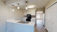 This single-story unit offers a practical and well-organized layout with a neutral color palette throughout. The kitchen serves as a central feature, designed with white cabinetry, white countertops, and a tile backsplash. Black appliances — including an electric range, built-in microwave, and dishwasher — provide a clean contrast against the bright cabinetry. A peninsula with a double-basin sink and pendant lighting above separates the kitchen from the adjacent living area, which flows openly and features wood-look flooring. The living space benefits from natural light filtering through blinds-equipped windows.

The bedrooms are carpeted and finished in warm neutral tones, each featuring windows that invite natural light. Ceiling fans with integrated lighting are present in multiple bedrooms, adding both function and comfort. At least one bedroom includes a walk-in closet with built-in shelving. The bathroom features a vanity with ample cabinet storage, a towel bar, and wood-look flooring. Textured ceilings are consistent throughout the unit, and the overall design emphasizes clean lines and a move-in-ready aesthetic.