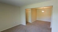 This single-floor residence offers a straightforward and functional layout designed for comfortable everyday living. The main living area is finished with wall-to-wall carpet in a warm neutral tone, creating a cohesive and inviting atmosphere throughout the space. Walls are painted in a soft, light beige, contributing to a bright and airy feel that makes the rooms appear open and welcoming.

A defining feature of the layout is the open passageway connecting the main living area to an adjoining room, which enhances the sense of flow and spaciousness between spaces. The adjoining area is illuminated by a pendant light fixture hanging from the ceiling, adding a subtle decorative touch while providing focused overhead lighting.

A louvered bi-fold closet door is visible within the connected space, offering convenient built-in storage. White baseboards run throughout, providing a clean contrast against the neutral walls and carpet. The overall design is understated and move-in ready, with a simple aesthetic that provides a versatile backdrop for a variety of furnishing styles. The single-floor plan ensures easy accessibility and seamless navigation throughout the home.