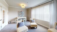 The Building Lounge at 675 Tea St in Bound Brook, NJ offers residents a thoughtfully designed communal gathering space finished with refined details and a cohesive contemporary aesthetic. Crown molding lines the ceilings throughout, adding an elegant architectural touch that elevates the overall ambiance of the room.

The lounge is organized into distinct functional zones. A comfortable seating area features a collection of yellow-and-white patterned accent chairs arranged around a white oval coffee table, creating an inviting space for relaxation or conversation. A wall-mounted flat-screen television anchors one side of the room, making it well-suited for group viewing. A set of large windows fitted with vertical blinds and sheer curtains floods the space with natural light while offering views of the surrounding greenery.

A dedicated dining nook provides a white high-top table accompanied by matching bar-height stools, ideal for casual meals or collaborative work sessions. A large decorative wall clock serves as a striking focal point in this area. Glass-paned French doors connect to an adjoining room, enhancing the sense of openness and flow.

The décor throughout is cohesive and modern, with neutral gray walls punctuated by gold-toned abstract artwork and warm-toned drum flush-mount light fixtures. Soft carpeting underfoot and carefully chosen decorative accents complete the welcoming, community-focused atmosphere of this shared lounge.