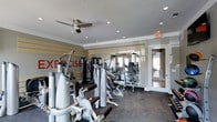 This fitness center is thoughtfully designed to support a comprehensive workout experience. The space is outfitted with a robust selection of strength-training machines, including cable systems, weight benches, and multi-function resistance equipment, all arranged to maximize floor space and ease of movement between stations. The flooring features a dark, durable surface well-suited to heavy equipment and high-traffic use. Overhead, recessed lighting is complemented by a ceiling fan, ensuring the space remains well-lit and comfortable during workouts. A large wall mirror spans a significant portion of the room, visually expanding the space and allowing users to monitor their form. Bold red lettering reading "EXERCISE" accents the wall, reinforcing the room's energetic and motivational atmosphere. The neutral color palette of grey and white throughout the walls and ceiling lends a clean, modern aesthetic. A glass-paneled door leads to an adjacent room, offering a glimpse of additional amenity space beyond. The overall design balances functionality with a polished, contemporary feel, making it a well-appointed facility for fitness enthusiasts.