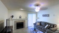 This single-story space centers around a well-appointed living room designed for comfort and everyday functionality. The room is anchored by a tile-surround fireplace with a clean white mantel, serving as a natural focal point and adding a warm, inviting atmosphere. A textured popcorn ceiling and ceiling fan with a light fixture provide both character and practical ventilation and illumination throughout the space.

Natural light enters generously through a sliding glass door dressed with vertical blinds, brightening the interior and offering the option for outdoor access. The living area comfortably accommodates plush seating, with ample space for a sofa arrangement and a central coffee table, making it well-suited for relaxing or entertaining.

Wall-to-wall carpeting adds softness underfoot, complementing the neutral, light-toned walls that keep the space feeling open and airy. Decorative wall art and subtle accent pieces contribute to a lived-in, homey aesthetic. Overall, the living area balances practical features with a cozy ambiance, highlighted by the fireplace as its defining architectural element.