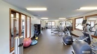 This fitness center is a comprehensively equipped single-floor facility designed to support a full range of workout routines. The space is organized into distinct functional zones, beginning with a dedicated cardio area that houses multiple treadmills and elliptical trainers, including Precor brand machines. Wood-framed windows along the walls allow natural light to filter in and provide outdoor views of surrounding greenery, creating an open and energizing atmosphere. Flush fluorescent lighting supplements natural light throughout the space.

The strength training zone features a variety of cable machines, weight benches, and multi-functional cable stations suitable for both isolated and compound exercises. A well-stocked dumbbell rack holds an extensive range of free weights, and a dedicated area accommodates medicine balls, stability balls, and kettlebells for functional and dynamic training. Large mirrors are strategically positioned along the walls to assist with form and to visually expand the space.

The flooring transitions between rubberized fitness surfaces in the weight and cardio areas, providing durability and safety. A beverage vending machine offers convenient refreshment access. The facility's neutral color palette, wood-trimmed windows, and clean layout contribute to a practical yet welcoming training environment.