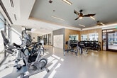 Located in Brookhaven, GA, this well-appointed two-level fitness center offers a comprehensive workout environment designed with both functionality and aesthetics in mind. The expansive main floor is bathed in natural light streaming through floor-to-ceiling glass windows, which also provide open views of the surrounding neighborhood. The layout accommodates a wide range of training modalities, including a dedicated cardio zone equipped with Matrix treadmills, ellipticals, and stationary bikes, as well as a strength training area featuring a full dumbbell rack, weight benches, and cable machines. Warm wood-paneled accent walls and large mirrors complement the polished concrete-style flooring, while ceiling fans and sleek linear lighting fixtures contribute to a clean, modern ambiance. A separate functional training room features a striking abstract mural, and is outfitted with kettlebells, battle ropes, resistance bands, medicine balls, TRX suspension trainers, and a multi-use rig — ideal for dynamic and cross-training workouts. Lockers are conveniently positioned within the main floor area. The upper mezzanine level, accessible via a staircase with a modern metal railing, offers additional equipment including rowing machines and specialty strength machines, along with bright windows overlooking the exterior.