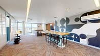 Located at 300 Massachusetts Ave in Amherst, Massachusetts, this contemporary communal lounge offers a well-appointed, open-plan gathering space designed with both comfort and creativity in mind. The room is anchored by striking bold geometric murals — one rendered in black and white, the other in deep navy — that serve as dramatic focal points and lend the space a distinctly modern, artistic character.

The lounge is organized into two functional zones. A relaxed seating area features a large, L-shaped sectional sofa in soft grey, paired with natural wood coffee tables and complementary accent chairs, ideal for casual conversation or unwinding. Adjacent to this is a dining and work area centered around a butcher-block high-top table with black bar stools, perfect for collaborative work or social gatherings.

A warm terracotta feature wall hosts a large flat-screen television flanked by built-in wood shelving and storage units, adding both functionality and a pop of color. Curated framed artwork lines one wall, contributing to the space's gallery-like atmosphere.

Throughout, a herringbone-patterned carpet in earthy, multi-toned hues adds warmth and texture underfoot. Recessed cove lighting along the ceiling creates a soft, ambient glow. Floor-to-ceiling windows flood the space with natural light and frame expansive views of the surrounding Amherst skyline, making this lounge as visually engaging as it is inviting.