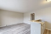 Located at 3203 Orange St in Greensboro, NC, this one-bedroom, one-bathroom unit offers a practical and efficient single-floor layout. The combined kitchen and living area forms the heart of the space, featuring light wood-style vinyl flooring, neutral-toned walls, and a textured ceiling. The kitchen is defined by a peninsula counter with a rounded edge that opens to the living area, creating an open, connected feel. It includes white cabinetry, a black electric range, a range hood, a refrigerator, and a sink with granite-style countertops. A baseboard heating unit runs along the living room wall, providing supplemental warmth. The bedroom is a generously sized retreat with plush gray carpeting, a ceiling fan with light fixture, a window with blinds that allows natural light, and a wall-mounted air conditioning unit. Two doorways lead from the bedroom — one to the walk-in closet, which is outfitted with wire shelving and hanging rods along two walls, and one back to the main living area. The bathroom features a clean, all-white tub and shower surround with a built-in safety grab bar. Throughout the unit, the clean, neutral palette and functional finishes create a straightforward and livable atmosphere.