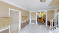 This single-story building features a neatly presented common area that serves as the central hub for residents. The entry lobby is finished with white tile flooring and two-tone walls — a warm tan upper half paired with a deeper brown wainscoting below — all framed by crisp white crown molding and pilasters that lend a classic, structured aesthetic. A wall-mounted bank of individual mailboxes provides a convenient mail collection point directly within the lobby area, adding to its functional design. An elegant arched passage connects the lobby to a long, carpeted corridor that extends deep into the building, lined on both sides by multiple unit doors evenly spaced along its length. The hallway is warmly lit overhead, creating a welcoming and secure feel throughout. A runner of dark carpet runs the length of the corridor, contrasting with the lighter tiled lobby. A flush-mount ceiling fixture in the lobby adds a touch of traditional detail to the overall interior. The space is clean, organized, and thoughtfully maintained, reflecting a well-managed residential building designed for both practicality and comfort.