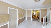 This single-story building features a neatly presented common area that serves as the central hub for residents. The entry lobby is finished with white tile flooring and two-tone walls — a warm tan upper half paired with a deeper brown wainscoting below — all framed by crisp white crown molding and pilasters that lend a classic, structured aesthetic. A wall-mounted bank of individual mailboxes provides a convenient mail collection point directly within the lobby area, adding to its functional design. An elegant arched passage connects the lobby to a long, carpeted corridor that extends deep into the building, lined on both sides by multiple unit doors evenly spaced along its length. The hallway is warmly lit overhead, creating a welcoming and secure feel throughout. A runner of dark carpet runs the length of the corridor, contrasting with the lighter tiled lobby. A flush-mount ceiling fixture in the lobby adds a touch of traditional detail to the overall interior. The space is clean, organized, and thoughtfully maintained, reflecting a well-managed residential building designed for both practicality and comfort.