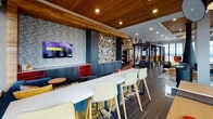 Located on an upper floor of 90 New St in New Brunswick, NJ, the Club Room is a thoughtfully designed amenity space that blends urban sophistication with creative, eclectic styling. Floor-to-ceiling windows wrap around multiple seating zones, flooding the space with natural light and offering sweeping panoramic views of the New Brunswick skyline and surrounding landscape.

The interior design is bold and distinctive, anchored by warm wood-paneled ceilings with recessed lighting that contrast beautifully against the dark tile flooring. A striking feature wall composed of a vibrant mosaic of colorful tiles serves as an artistic focal point, complemented by a large flat-screen television and dark custom cabinetry with open shelving accented in red.

Seating throughout the space is diverse and inviting, ranging from a long communal counter with modern bar-height chairs ideal for working or casual dining, to cozy lounge clusters featuring accent chairs in vivid hues. Corner seating vignettes with bistro tables positioned directly against the expansive windows offer intimate spots to take in the city views.

Geometric patterned area rugs, bold graphic murals, and carefully curated décor details add layers of visual energy throughout. The overall ambiance is vibrant yet comfortable, making the Club Room a dynamic gathering space suited for both social events and quiet retreat.