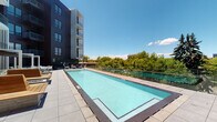This contemporary outdoor amenity level offers a thoughtfully designed rooftop experience centered around a large, rectangular lap-style pool with pristine turquoise water and clean tile surroundings. The pool deck is framed by glass railings that preserve open sightlines to the surrounding treetop and city views under expansive blue skies.

Adjacent to the pool, a spacious outdoor terrace provides multiple distinct zones for relaxation and entertainment. A pergola structure with a slatted overhead canopy offers partial shade over a well-appointed lounge area furnished with modern white-framed sofas and chairs accented with bold red and patterned cushions. The seating areas are arranged to encourage both social gatherings and quiet relaxation.

An outdoor kitchen station features a stainless steel gas grill set within warm wood cabinetry, ideal for al fresco dining. Decorative stone planters with black gravel and low-profile greenery add a polished, landscaped touch throughout the terrace.

Large floor-to-ceiling sliding glass doors connect the outdoor deck to an interior lounge space, which is visible through the glass and features warm lighting, a large-screen television, patterned tile flooring, and comfortable seating — creating a seamless indoor-outdoor flow. String lights add an inviting ambiance to the transitional space, making it equally functional day or night.