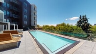 This contemporary outdoor amenity level offers a thoughtfully designed rooftop experience centered around a large, rectangular lap-style pool with pristine turquoise water and clean tile surroundings. The pool deck is framed by glass railings that preserve open sightlines to the surrounding treetop and city views under expansive blue skies.

Adjacent to the pool, a spacious outdoor terrace provides multiple distinct zones for relaxation and entertainment. A pergola structure with a slatted overhead canopy offers partial shade over a well-appointed lounge area furnished with modern white-framed sofas and chairs accented with bold red and patterned cushions. The seating areas are arranged to encourage both social gatherings and quiet relaxation.

An outdoor kitchen station features a stainless steel gas grill set within warm wood cabinetry, ideal for al fresco dining. Decorative stone planters with black gravel and low-profile greenery add a polished, landscaped touch throughout the terrace.

Large floor-to-ceiling sliding glass doors connect the outdoor deck to an interior lounge space, which is visible through the glass and features warm lighting, a large-screen television, patterned tile flooring, and comfortable seating — creating a seamless indoor-outdoor flow. String lights add an inviting ambiance to the transitional space, making it equally functional day or night.