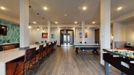 This single-floor community clubroom is designed for social gathering and recreation, offering a well-appointed layout that blends lounge, dining, and game-room functions seamlessly. The open floor plan is anchored by large-format tile flooring in a wood-look finish, which runs throughout the space and contributes to its contemporary aesthetic. Structural columns define distinct activity zones without interrupting the room's spacious, connected feel.

A long bar-height communal table with upholstered stools runs along one side of the room, ideal for group gatherings or casual dining. Pendant lighting overhead adds a warm, inviting ambiance to this area. The game zone features both an air hockey table and a foosball table, providing ample entertainment options for residents.

A dedicated wet bar area stands out as a design highlight, showcasing striking teal elongated hexagonal tile as a full backsplash, navy cabinetry with matte hardware, a stainless steel undermount sink, and a wall-mounted television above — making it a natural focal point for entertaining.

Recessed lighting throughout ensures the space is well-lit, while wall sconces in the lounge seating nook add warmth. Colorful local artwork adorns the neutral gray walls. Multiple sets of glass double doors provide access to an adjacent outdoor area, flooding the room with natural light and creating a seamless indoor-outdoor connection.