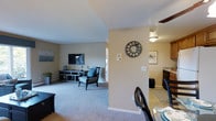 This single-floor residence features 2 bedrooms and 2 bathrooms, thoughtfully arranged to maximize functionality and comfort. The open layout seamlessly connects the living, dining, and kitchen areas, creating a cohesive flow throughout the main living space. The living room is carpeted and offers ample room for seating and entertainment, highlighted by a media console and a decorative accent chair with blue cushioning, adding a touch of color to the neutral palette. Warm beige walls and flush-mount ceiling lighting provide a welcoming ambiance throughout. A partial wall divides the living area from the dining space, offering a sense of definition without sacrificing openness. The dining area accommodates a table and chairs and transitions naturally into the kitchen, which features tile flooring and dark cabinetry visible in the background. Decorative touches, including a sunburst wall clock and framed artwork, add personality to the space. A ceiling fan is also present, contributing to year-round comfort. The overall design emphasizes a clean, neutral aesthetic that is both practical and inviting.