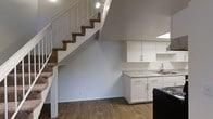 This two-story residence offers a well-organized layout across two levels, designed to maximize both functionality and comfort. The main floor showcases durable hardwood-style flooring that runs throughout the open living area, creating a clean and cohesive aesthetic. The kitchen is outfitted with bright white cabinetry featuring brushed nickel hardware, light granite-style countertops, and a stainless steel sink, all illuminated by natural light from a skylight above. Dark appliances provide a subtle contrast to the crisp white cabinetry. A carpeted staircase with a white-railed banister rises from the main level to the upper floor, adding a classic touch to the interior. Recessed and track lighting throughout the main level ensures the space is well-lit, enhancing the clean, neutral color palette of soft gray walls and white trim. The open design of the main floor allows for versatile furniture arrangements, while the two-story layout provides a natural separation between living and private spaces. Overall, the residence presents a tidy, move-in-ready environment with practical finishes and a straightforward, livable design.