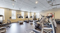 Located at 1982 State Highway 70 E in Cherry Hill, NJ, this fitness center offers a well-appointed exercise environment designed to support a full range of workout routines. The space is laid out on a single floor and provides ample room for both cardio and strength training activities.

The interior features a drop-panel ceiling with recessed lighting and ceiling fans, ensuring consistent airflow and visibility throughout. Warm, neutral-toned walls are complemented by large windows fitted with white shutters, allowing natural light to fill the space and offering views of the surrounding greenery outside.

Cardio equipment is prominently featured, including multiple treadmills, elliptical trainers, and stationary bikes arranged to maximize usable floor space. Strength training is well supported with cable machine systems, a multi-function weight station, and a dedicated dumbbell rack. A large wall-mounted mirror along one section expands the sense of space and allows users to monitor their form during workouts.

Wall-mounted flat-screen televisions provide entertainment options during exercise sessions. Rubberized flooring throughout offers durability and safety underfoot. Lockers and seating are available near the entrance, and French doors lead to an adjacent area, enhancing the overall accessibility of the facility. Framed artwork adds a finishing touch to the clean, functional aesthetic of this fitness space.