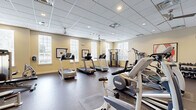 Located at 1982 State Highway 70 E in Cherry Hill, NJ, this fitness center offers a well-appointed exercise environment designed to support a full range of workout routines. The space is laid out on a single floor and provides ample room for both cardio and strength training activities.

The interior features a drop-panel ceiling with recessed lighting and ceiling fans, ensuring consistent airflow and visibility throughout. Warm, neutral-toned walls are complemented by large windows fitted with white shutters, allowing natural light to fill the space and offering views of the surrounding greenery outside.

Cardio equipment is prominently featured, including multiple treadmills, elliptical trainers, and stationary bikes arranged to maximize usable floor space. Strength training is well supported with cable machine systems, a multi-function weight station, and a dedicated dumbbell rack. A large wall-mounted mirror along one section expands the sense of space and allows users to monitor their form during workouts.

Wall-mounted flat-screen televisions provide entertainment options during exercise sessions. Rubberized flooring throughout offers durability and safety underfoot. Lockers and seating are available near the entrance, and French doors lead to an adjacent area, enhancing the overall accessibility of the facility. Framed artwork adds a finishing touch to the clean, functional aesthetic of this fitness space.