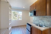 Located at 8525 163rd Ct NE in Redmond, Washington, this efficient studio unit offers a thoughtfully designed single-floor layout that maximizes every square foot. The open-plan living, dining, and kitchen area is unified by warm-toned laminate wood flooring that runs throughout, creating a cohesive and welcoming atmosphere. A large window floods the main living space with abundant natural light and frames views of the surrounding greenery and neighboring architecture.

The kitchenette is a functional highlight, featuring rich warm-toned wood cabinetry, granite-style countertops with a stone backsplash, a stainless steel sink, a compact refrigerator, and a microwave unit mounted above the counter — providing all essential amenities in a space-conscious footprint.

Ample built-in shelving and wire rack storage units line one wall of the living area, offering generous organizational options that are rarely found in similarly sized units. An additional shelving unit is positioned near a secondary window, which brings in further natural light and outdoor views of mature trees.

The bathroom is clean and straightforward, featuring a walk-in shower with white tile surround. Neutral wall tones throughout the unit contribute to a bright, airy ambiance. This unit is well-suited for those seeking a practical and efficiently organized living space in the heart of Redmond.