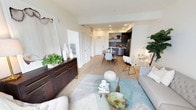 This single-floor unit showcases a thoughtfully designed open-concept layout that integrates the living, dining, and kitchen areas into one cohesive, flowing space. Light-toned hardwood flooring runs throughout, contributing to a bright and airy atmosphere that is further enhanced by recessed lighting along the ceiling.

The living area is anchored by a glass-topped coffee table and neutral upholstered seating, creating a welcoming and comfortable space. A dark wood credenza with gold hardware adds a bold contrast against the soft, cream-toned walls, providing both storage and a stylish focal point.

The dining area sits centrally within the open floor plan, featuring a wooden dining table paired with modern white chairs, comfortably accommodating several guests. Greenery and decorative accents throughout the space add warmth and a lived-in character.

The kitchen is finished with dark cabinetry, stone tile backsplash, stainless steel appliances including a refrigerator and dishwasher, and offers functional counter space. The seamless transition between areas makes the overall layout feel spacious and well-connected, ideal for both daily living and entertaining.