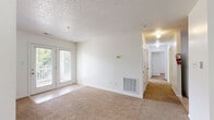 Located at 100 Ridgewood Ln in Radford, Virginia, this single-floor residence offers two bedrooms, two bathrooms, and a practical, well-organized layout. The living area is carpeted in warm beige tones beneath a textured ceiling, and French doors provide natural light while opening to an outdoor balcony with views of the surrounding greenery. A pass-through window connects the living space to the kitchen, which features warm oak cabinetry, light countertops, white appliances including a range and refrigerator, and tile flooring — creating an open, communicative flow between the two spaces. The primary bedroom is a comfortable, well-proportioned room with carpeted floors, a natural light window, and closet access. The second bedroom mirrors this layout with its own window and carpeted floor. Three closets throughout the unit provide ample storage. Both bathrooms are finished with tile flooring; one includes a tub-shower combination, while the other features a walk-in shower, a vanity with oak cabinetry, and a lighted mirror. The overall aesthetic is clean and neutral, with white walls and consistent finishes throughout, offering a move-in-ready space in a convenient Radford location.