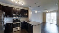 This single-story space showcases a well-appointed kitchen as its centerpiece, designed with a clean, contemporary aesthetic. The kitchen features rich, dark espresso cabinetry that extends to the upper walls, providing ample storage while creating a bold visual contrast against the neutral-toned walls. Granite countertops with subtle veining run throughout, including across a peninsula that doubles as a breakfast bar, offering additional workspace and casual dining options.

Stainless steel appliances, including a range, microwave, and dishwasher, lend a polished, professional feel to the cooking area. A stainless undermount sink is positioned in the peninsula for added convenience. Track lighting above the main counter and elegant pendant lights over the peninsula ensure thorough, layered illumination throughout the kitchen.

Crown molding adds a refined architectural detail along the ceiling line, enhancing the overall sophistication of the space. Dark hardwood flooring flows seamlessly from the kitchen into the adjoining open living area, creating a cohesive and connected layout. The transition between these areas feels natural and spacious, making the overall space ideal for both everyday living and entertaining.