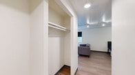 This efficiently designed single-floor unit offers a well-organized layout across four distinct areas, making smart use of its compact footprint. Light wood-tone flooring runs throughout the space, providing a warm and cohesive aesthetic.

The galley-style kitchen is outfitted with grey cabinetry, white countertops, a stainless steel refrigerator, and a built-in range with sink — offering a fully functional cooking space despite its streamlined form.

The main living area features a large window dressed with blackout roller shades, allowing for natural light while maintaining privacy. A low-profile media console with open shelving and drawer storage adds practical organization to the room.

The sleeping area connects seamlessly to the living space and includes a built-in closet with a full-width hanging rod and shelf, offering ample wardrobe storage. A ceiling fan with integrated lighting provides both comfort and illumination.

The bathroom features a single-sink vanity with a white countertop, a large mirror, and distinctive light blue square wall tiles that add a classic, clean character to the space. Track lighting in the ceiling is visible from the bathroom, reinforcing the consistent, modern-utilitarian design found throughout the unit.