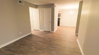 This single-floor residence offers a clean, open layout with a cohesive design that flows naturally from one area to the next. Rich, dark hardwood-style flooring runs throughout the main living space, providing a warm and grounded aesthetic that complements the neutral gray-toned walls. White baseboards and trim offer a crisp contrast, giving the interior a polished, well-maintained appearance. A textured ceiling adds subtle dimension overhead.

The main living area is open and spacious, with two white-paneled doors that likely lead to closets or utility spaces, offering practical built-in storage. The layout opens seamlessly toward a dining area, where an elegant chandelier serves as a refined focal point, adding a touch of sophistication to the space.

Beyond the dining area, the kitchen is visible and equipped with stainless steel appliances, including a microwave and a dark range, set against light cabinetry. The kitchen's compact yet functional design makes efficient use of the available space. Vent registers are positioned along the upper walls, indicating a central air and heating system throughout. Overall, the space presents a tidy, move-in-ready interior with consistent finishes and a welcoming atmosphere.