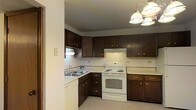 This single-floor residence offers a practical and straightforward layout designed for comfortable everyday living. The kitchen serves as a central feature of the home, showcasing abundant dark wood cabinetry that provides generous storage space both above and below the countertops. The kitchen is equipped with a white electric range with oven, an under-cabinet range hood, and a double-basin sink positioned at the corner with a standard faucet. A dishwasher is also integrated into the kitchen setup. The countertops are light-toned, offering a clean contrast against the rich, dark cabinetry. Overhead, a multi-bulb chandelier-style light fixture provides ample illumination throughout the kitchen space. The flooring in the kitchen area carries a neutral, low-pile carpet that extends into the adjoining areas, contributing to a cohesive and understated aesthetic throughout the home. The overall ambiance of the space is functional and classic, with a no-frills design that prioritizes utility and ease of use. The layout appears well-organized, making efficient use of the available space while maintaining a tidy and livable environment.