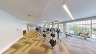 This expansive fitness center offers a comprehensive and modern exercise environment spread across a generously sized single-floor layout. The space is defined by its striking herringbone-patterned wood-look flooring, which adds a refined aesthetic throughout the room. Abundant natural light pours in through large floor-to-ceiling windows that line the exterior walls, offering views of a lush outdoor area featuring a pool and palm trees, creating an energizing atmosphere for workouts.

The facility is outfitted with a wide array of professional-grade equipment from brands such as Hoist and WaterRower, catering to a full spectrum of fitness needs. Cardio equipment includes treadmills, ellipticals, stationary bikes, and rowing machines, all thoughtfully arranged to maximize space and usability. The strength training section features multiple cable machines, selectorized weight machines, a Smith machine loaded with free weights, and additional resistance-based stations.

Recessed and linear LED lighting supplements the natural light, ensuring the space remains bright and well-lit at all times. Full-length mirrors along one wall enhance the sense of openness and allow users to monitor their form. The clean, neutral color palette of white walls and light tones throughout contributes to a modern, professional ambiance that is both functional and visually appealing.
